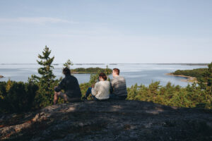 valokuva, kuvassa kesäinen maisema saaristosta, kuvakulma aavalle merelle, etualalla kallion päällä istuu kolme nuorta ihmistä selin