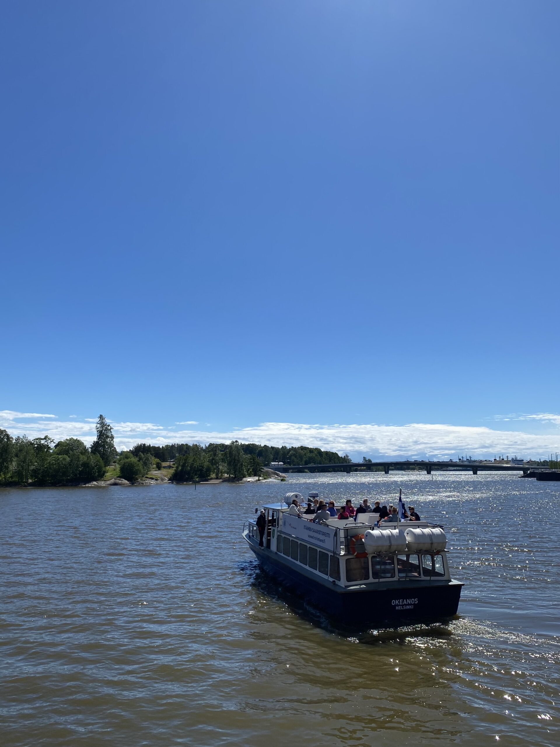 Vene, jonka kannella on ihmisiä ajamassa poispäin merelle. Taustalla näkyy metsäistä rantaa.