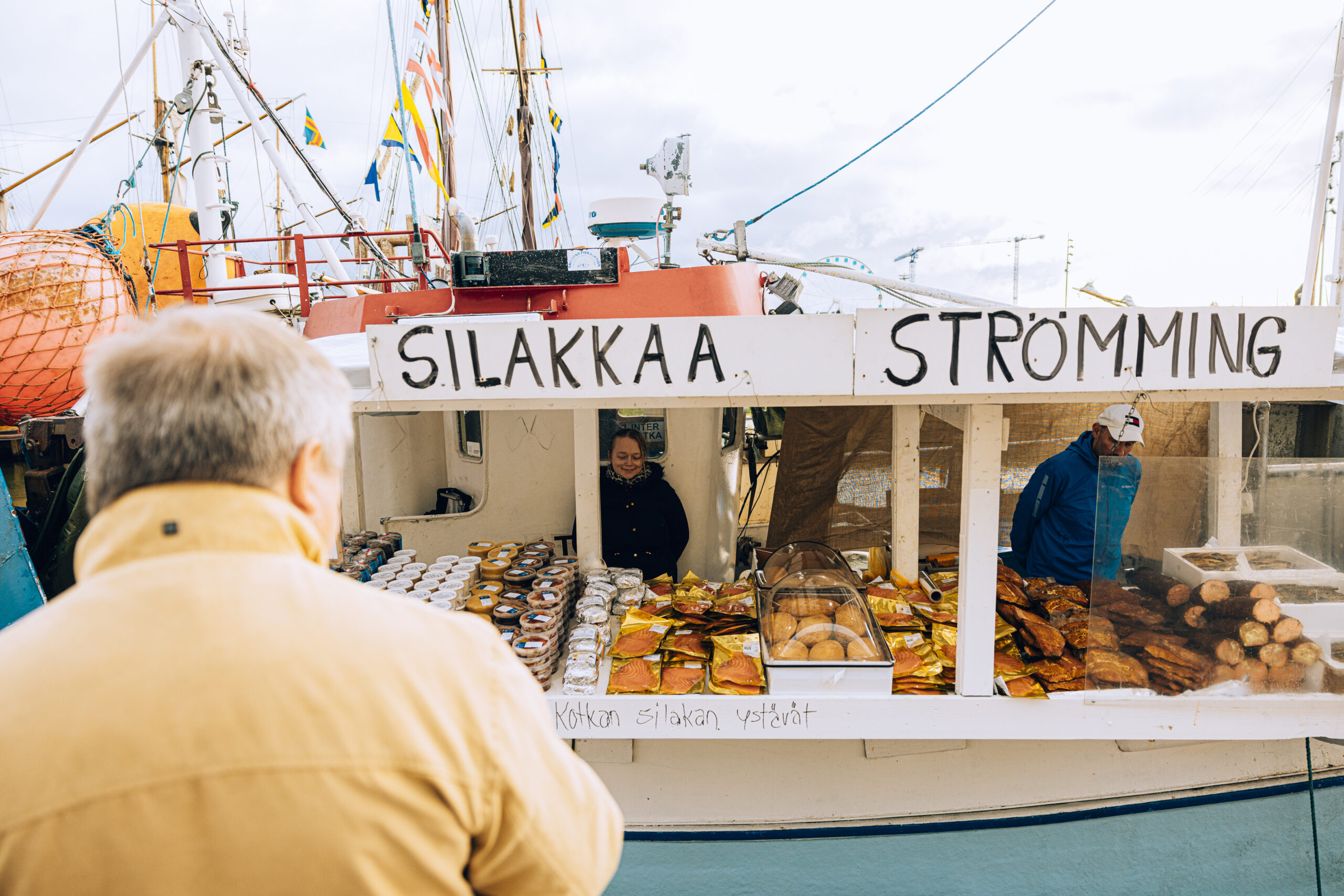 Koju, jossa lukee isolla Silakkaa Strömming. Kojussa on kalatuotteita ja sen takana myyjä. Etualalla henkilö selin keltaisessa takissa.