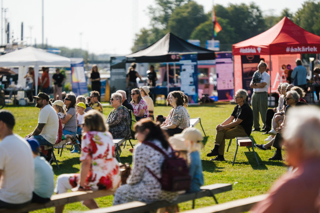 People sit on benches on a summer day and watch the show. In the background, exhibition tents on the lawn.