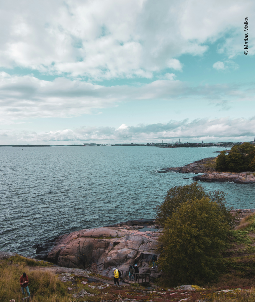 Ihmiset kulkevat kalliolla meren rannalla. Taivaalla pilvet repeilevät. Meri aaltoilee hieman. 