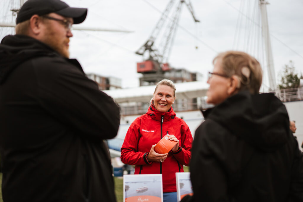 Punatakkinen henkilö juttelee kahdelle tumma-asuiselle henkilölle, jotka ovat selin kuvaan.